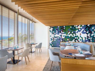 Café moderne avec plafond à poutres en bois, grandes fenêtres et mosaïque murale colorée au bord de la mer.