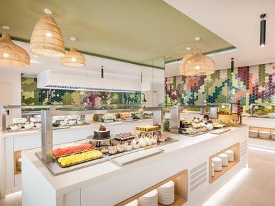 Modernes Buffet mit Obst, Backwaren und buntem Wandmosaik in hellen Farben und Holzdekor.