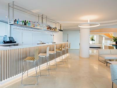 Espace bar d'hôtel moderne et lumineux avec des tabourets, un comptoir illuminé et des coins salon confortables.