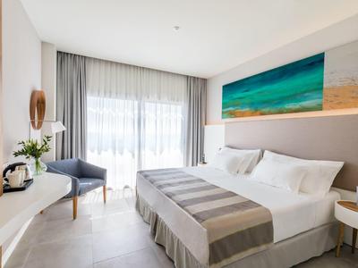 Helles Schlafzimmer mit großem Bett, grauen Vorhängen, blauem Sessel und Meerbild an der Wand.