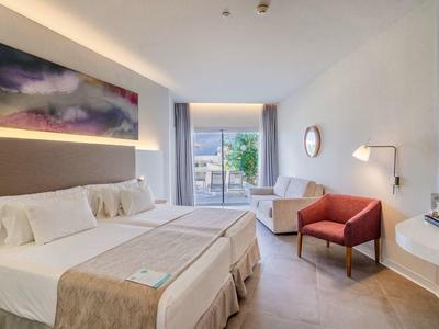 Hell eingerichtetes Hotelzimmer mit Doppelbett, rotem Sessel und Terrasse mit Blick ins Grüne.