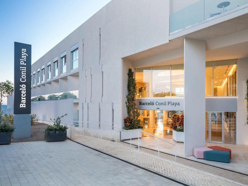 Moderner Hotelleingang mit Glasfront, weißen Wänden und Pflanzen in großen Töpfen.