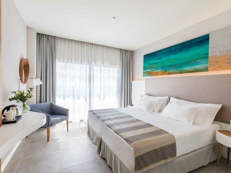 Helles Schlafzimmer mit großem Bett, grauen Vorhängen, blauem Sessel und Meerbild an der Wand.