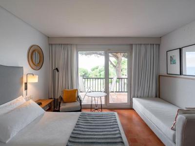 Nowoczesny pokój hotelowy z łóżkiem, sofą, krzesłem i balkonem z widokiem na zielone drzewa.