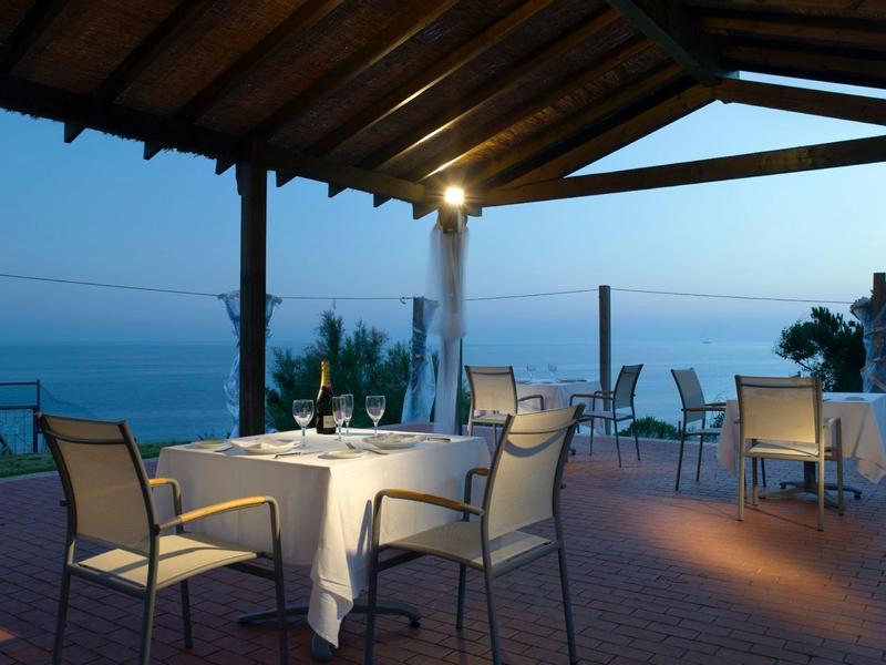 Abendliche Restaurantterrasse mit gedeckten Tischen und Meerblick unter einem Holzdach.
