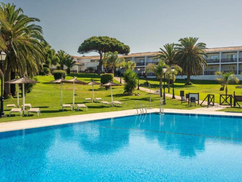 Großer Pool mit blauer Wasseroberfläche vor einem Hotel und grüner Gartenanlage.