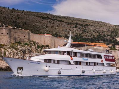 Nave passeggeri bianca su acque calme con collina boschiva e muro di pietra antico sullo sfondo.