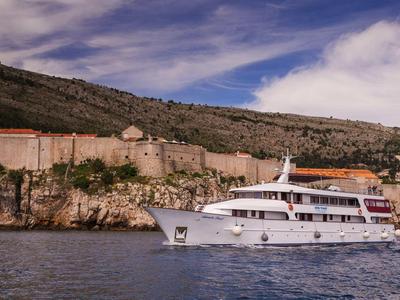 Yacht bianco che naviga vicino a una costa con mura e edifici storici sotto un cielo blu.