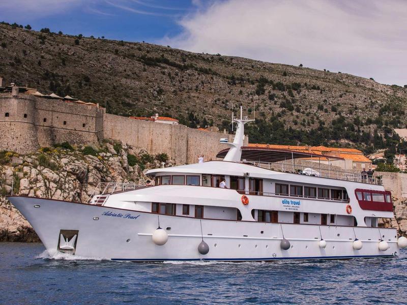 Nave passeggeri bianca su acque calme con collina boschiva e muro di pietra antico sullo sfondo.