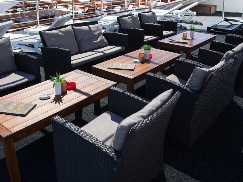 Area esterna con tavoli in legno e sedie imbottite su pavimento scuro, decorata con piccole piante.