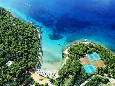 Luftaufnahme eines Strandes mit klarem blauem Wasser, umgeben von Wald und Sportplätzen.