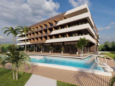 Modern hotel with pool and palm trees, surrounded by green landscape and blue sky.