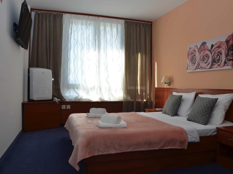 Camera d'hotel semplicemente arredata con letto matrimoniale, TV, frigorifero e finestra con tende.