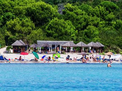 Beach with lounge chairs, umbrellas, and a building in front of a wooded hill.