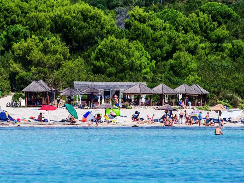 Strand mit vielen Menschen, Sonnenschirmen und kleinen Holzhütten vor grünem Wald.