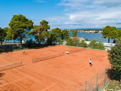 Terrain de tennis en terre battue rouge avec une personne, entouré d'arbres avec vue sur l'eau et le ciel.