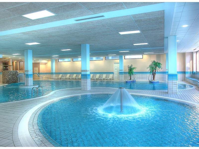 Grande piscine intérieure avec fontaine et espaces de détente modernes dans une zone bien-être lumineuse.