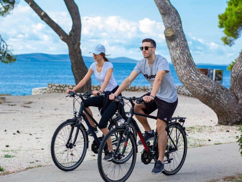 Un couple fait du vélo le long de la côte avec des arbres et la mer en arrière-plan.