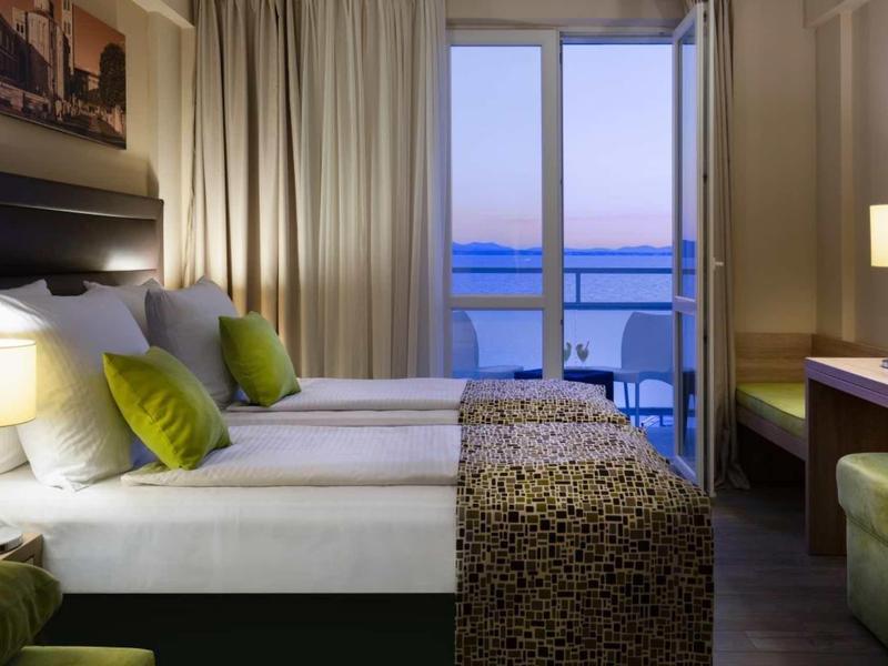 Chambre d'hôtel moderne avec deux lits, oreillers verts et vue sur la mer par de grandes fenêtres.