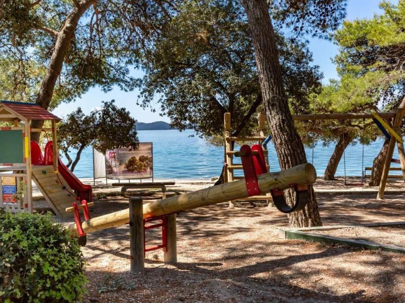 Aire de jeux avec balançoire à bascule et toboggan dans un espace boisé ombragé près de la mer.
