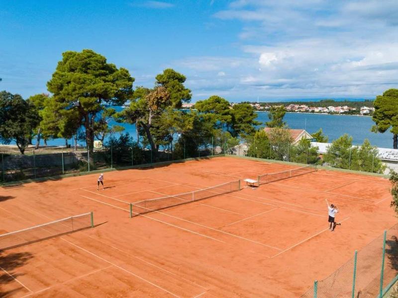 Terrain de tennis en terre battue rouge avec deux joueurs et vue sur la mer et les arbres.