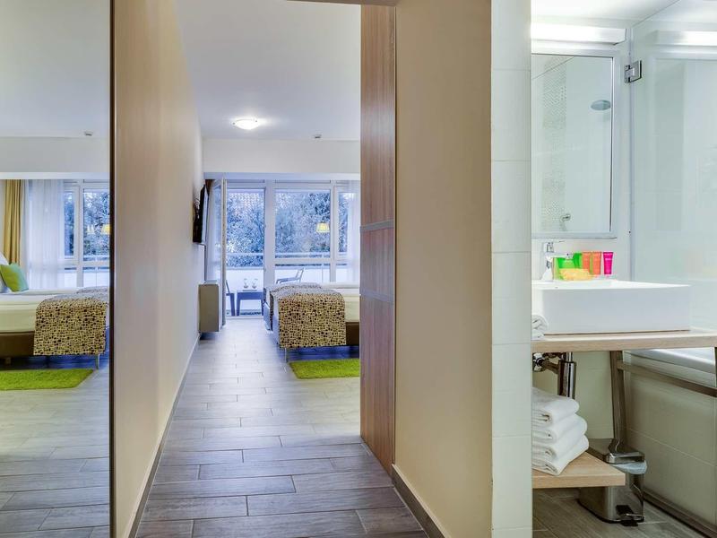 Vue à travers un couloir d'hôtel vers des chambres lumineuses avec lits et balcon, salle de bain avec lavabo à droite.