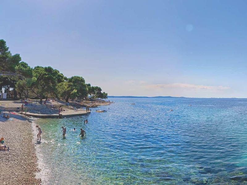 Petite plage de galets avec des arbres sur le bord et une mer bleu clair sous un ciel dégagé.