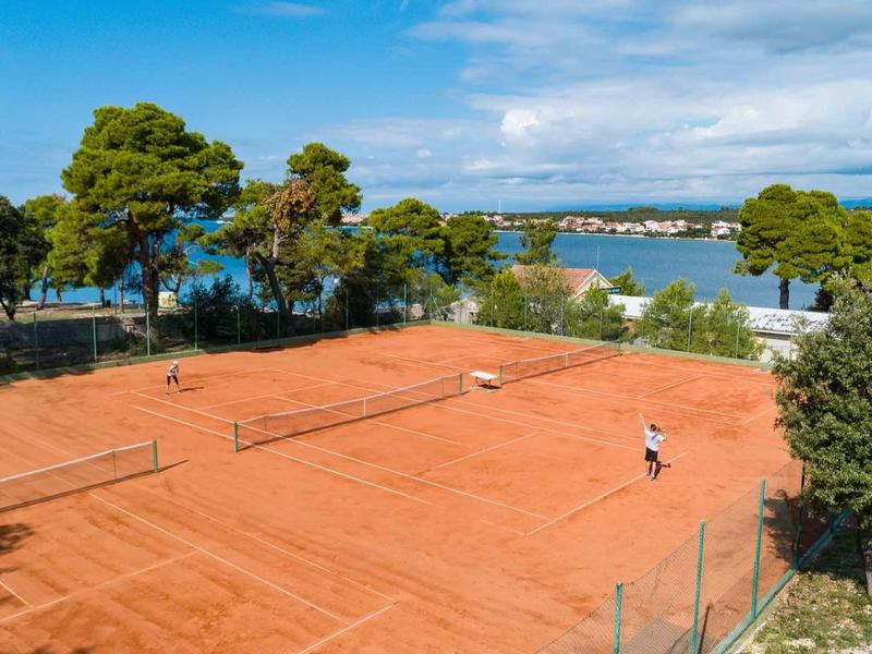 Terrain de tennis en terre battue rouge avec une personne, entouré d'arbres avec vue sur l'eau et le ciel.