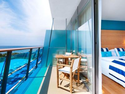 Balkon z szklaną balustradą, dwoma krzesłami i widokiem na morze obok nowoczesnej sypialni z niebiesko-białą pościelą.