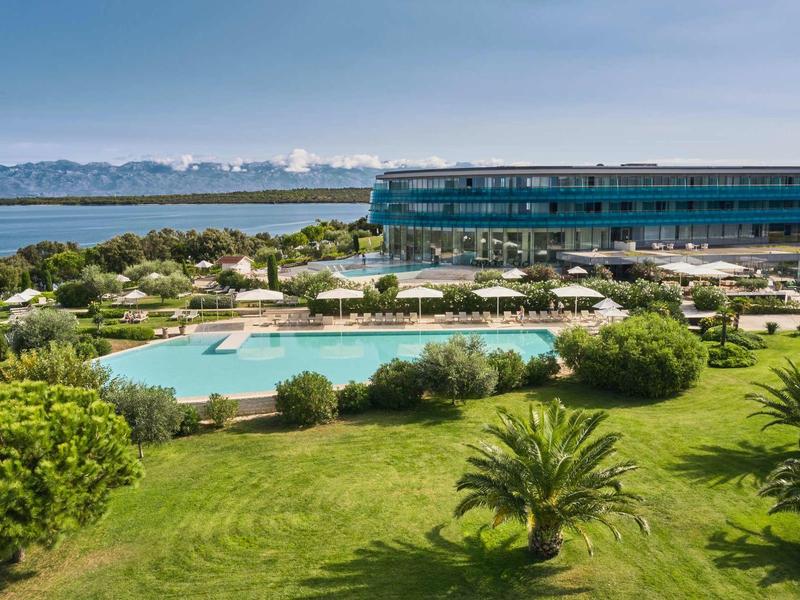 Widok na hotel z dużym basenem, zielonymi trawnikami i morzem w tle.