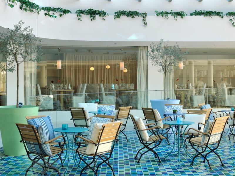 Taras z kolorowymi krzesłami i stołami przed dużymi oknami z roślinami.