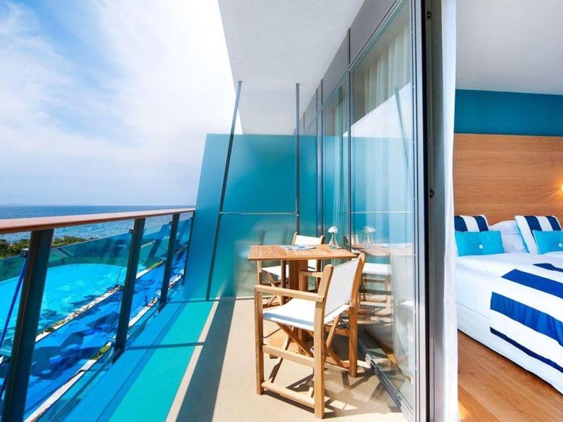 Balkon z szklaną balustradą, dwoma krzesłami i widokiem na morze obok nowoczesnej sypialni z niebiesko-białą pościelą.