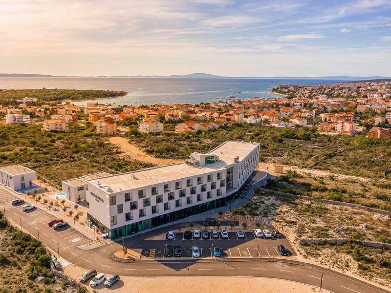 Modern hotelgebouw met parkeerplaats, omgeven door groen vlakbij de kust onder een blauwe hemel.
