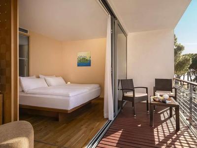 Camera d'hotel con balcone, tavolo, due sedie e vista su alberi e acqua.