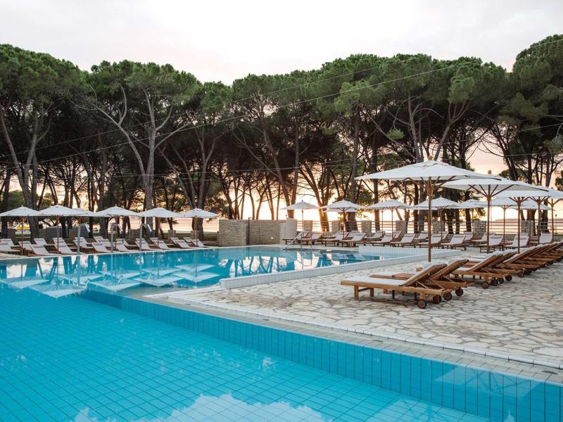 Una piscina blu con lettini e ombrelloni circondata da alberi al tramonto.