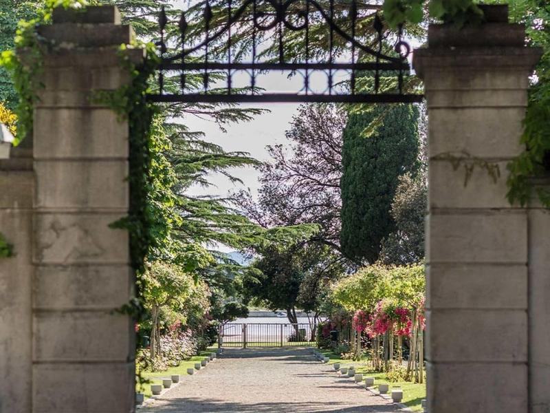 Un arco di pietra coperto di edera che conduce a un sentiero soleggiato nel giardino con panchine.