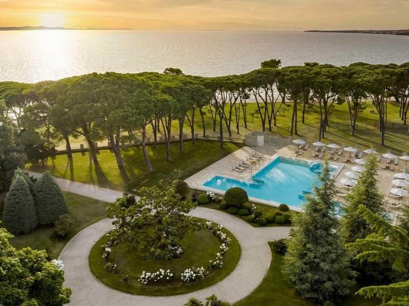 Complesso alberghiero con grande piscina, circondato da alberi e vista mare al tramonto