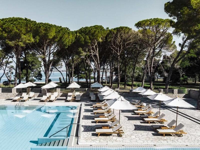 Piscina d'hotel moderna con lettini e ombrelloni davanti a una fila di alberi e vista mare.