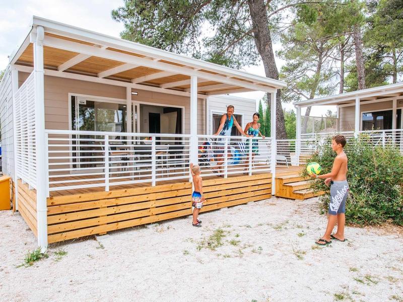 Family stands in front of a modern mobile home with a porch in a holiday park.