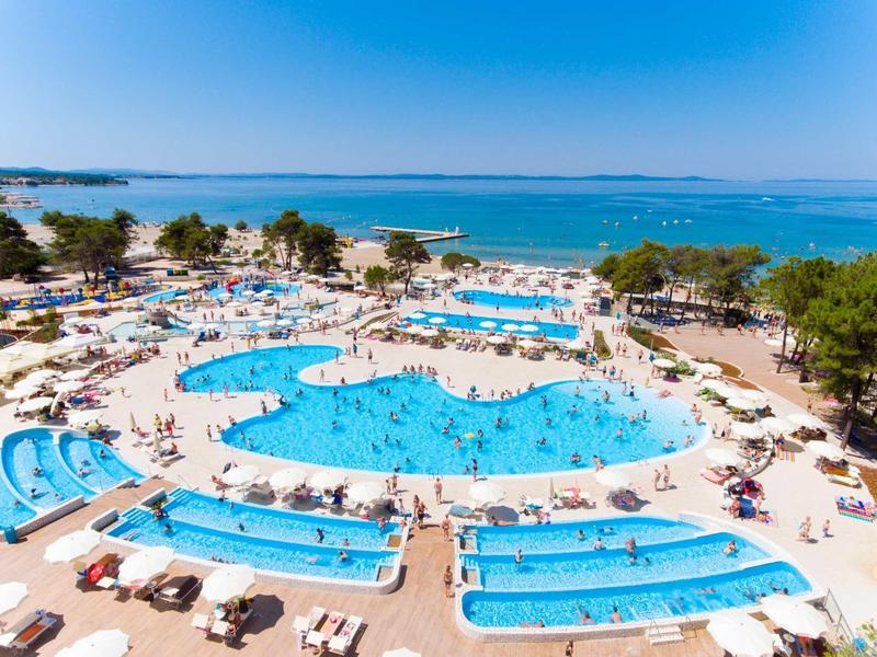 Large hotel pool with water slides, umbrellas, and people by the coast.