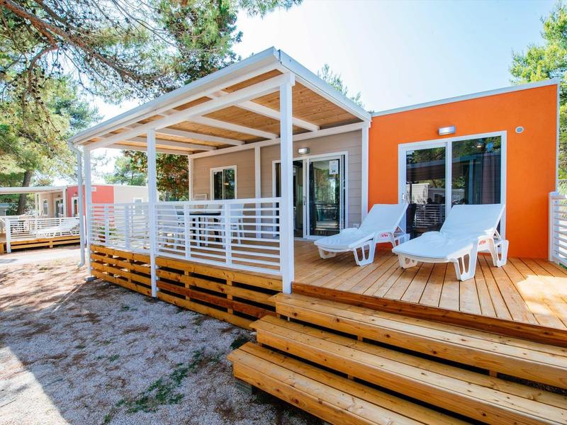 Modern holiday home with terrace, loungers, and surrounded by trees on sandy ground.