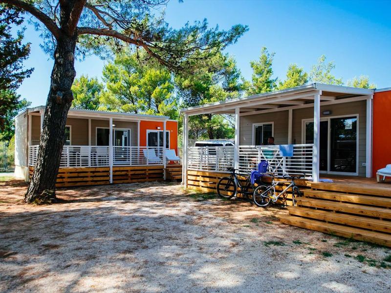 Two modern mobile homes with verandas in a sunny, wooded setting.