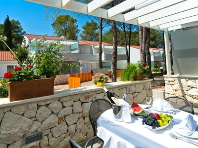 Terrasse mit weiß gedecktem Tisch, Blumen in Töpfen auf Steinmauer, dahinter Häuser und Bäume.
