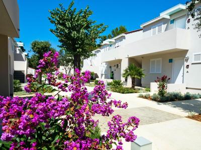 Hotel moderno con facciata bianca, sentiero e piante fiorite viola nel giardino sotto un cielo azzurro.