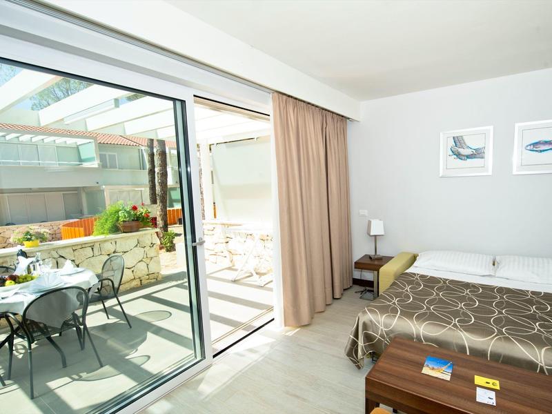 Jasny pokój hotelowy z łóżkiem podwójnym, szafką nocną i tarasem z krzesłami i stołem.