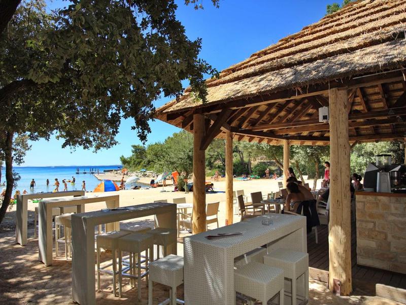 Strandbar mit Holzdach, weißen Tischen und Stühlen, umgeben von Bäumen und Sandstrand.