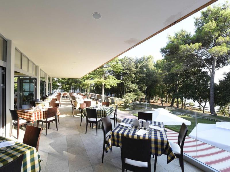 Eine helle Terrasse mit Tischen, Stühlen und karierten Tischtüchern, umgeben von Bäumen.