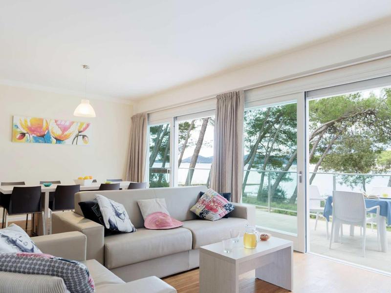 Helles Wohnzimmer mit grauem Sofa, Esstisch, bunten Kissen und großer Glasfront mit Blick auf Bäume.