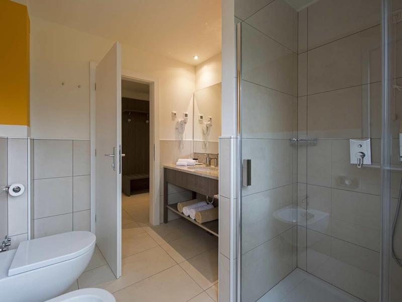 Helles Badezimmer mit Glasdusche, weißem WC, Doppelwaschbecken und großen weißen Fliesen.