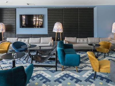 Salon d'hôtel moderne avec fauteuils bleu et jaune, canapé gris et tapis décoratif.